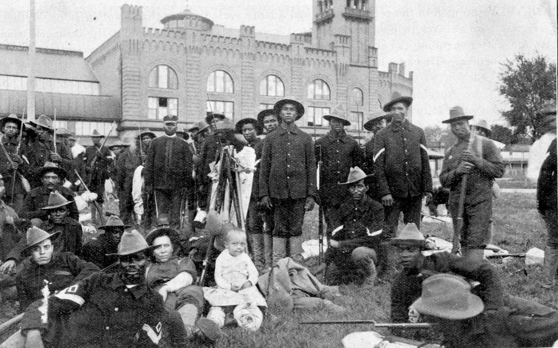 Approximately 30 Black men in hats and uniform, standing, sitting, and laying outside holding guns.