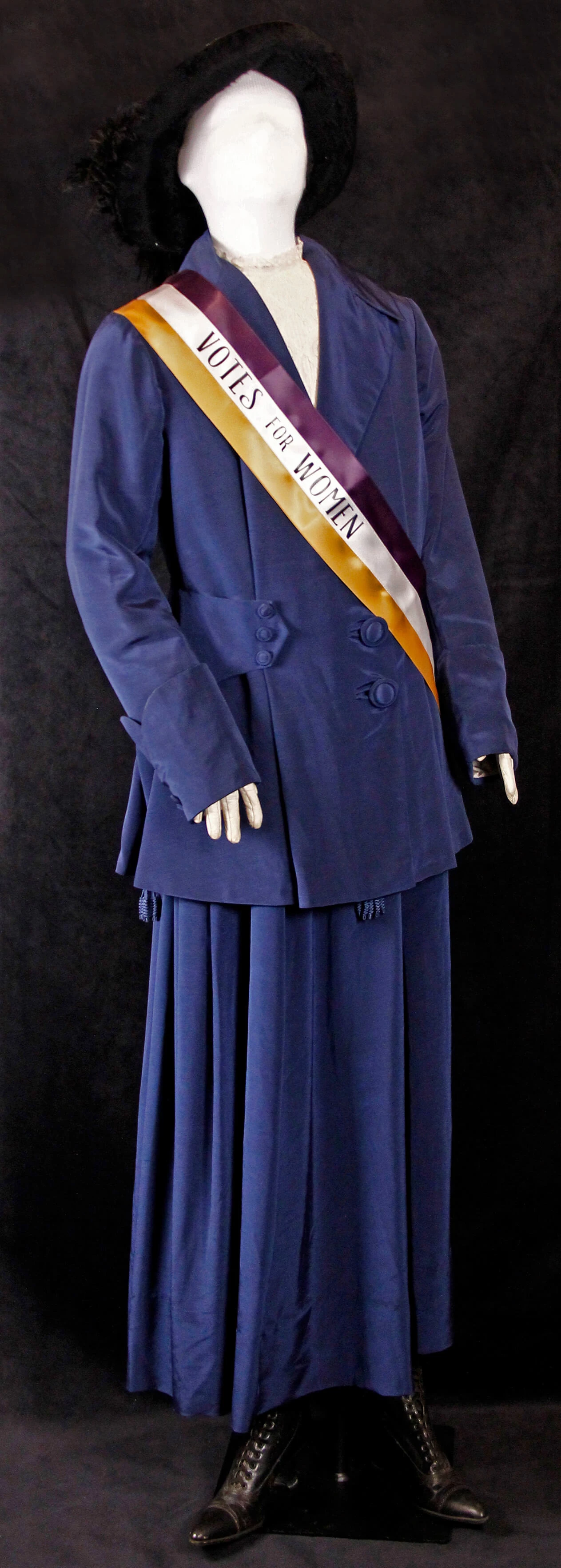 Mannequin dressed in blue velvet women's skirt and jacket, and black hat. A yellow and purple sash reads 'Votes for Women'