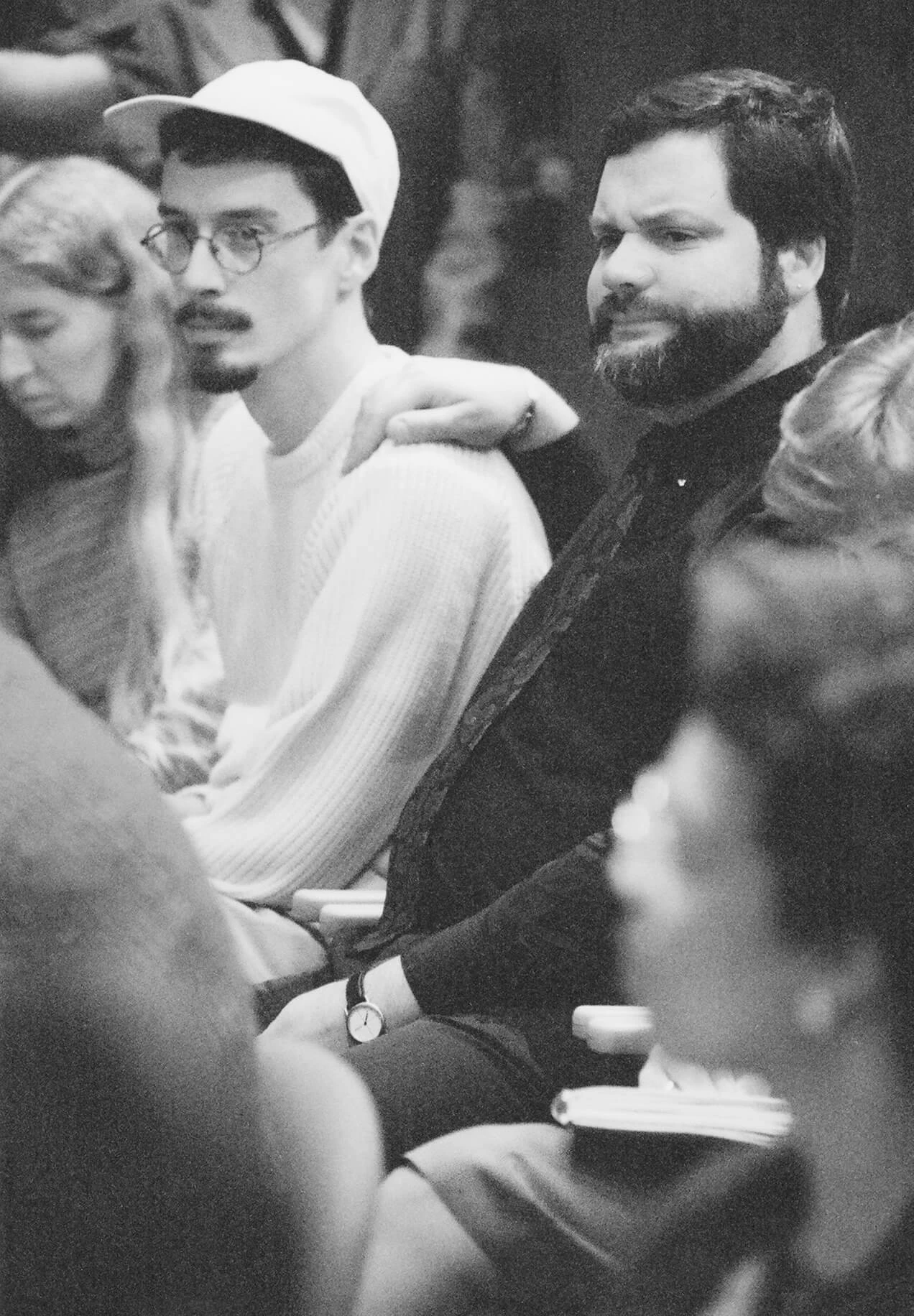 Black and white photo of two white men, one with a dark goatee wearing a light shirt and a light colored ball cap, the other wearing  a dark button up shirt with a dark beard.