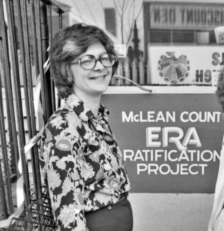 A white woman with glasses, short dark hair with light patches, a floral button up shirt stands on a staircase. behind her you can see part of a sign that reads 