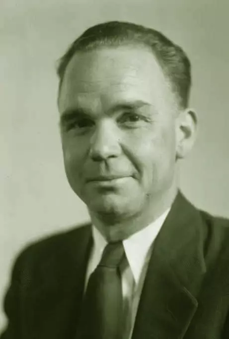 A light-skinned man with dark hair looks into the camera with a closed-mouth, one-sided smile. He is wearing a suit and tie.