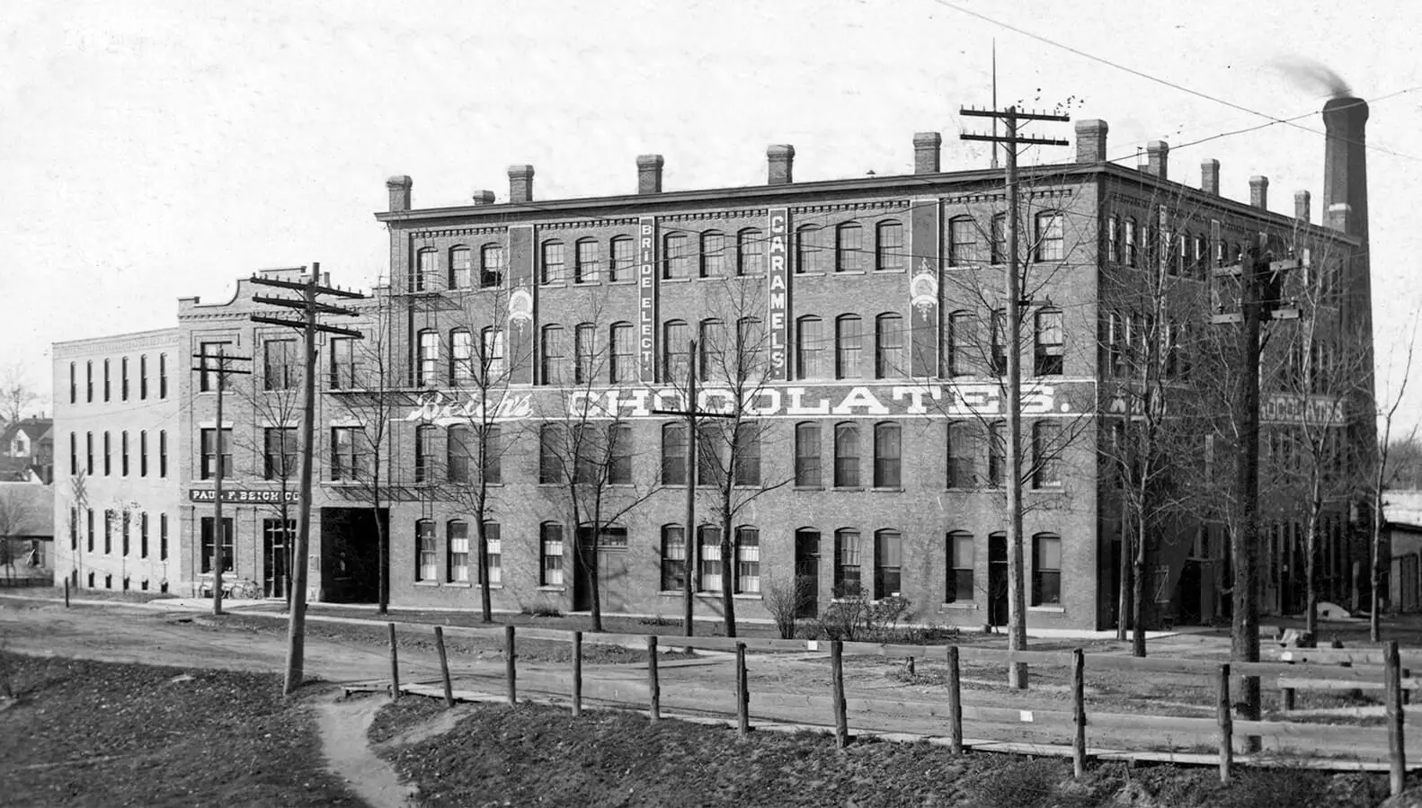 Large 4-story brick factory building. Between the 2nd and 3rd story it says beich chocolates and written vertically on the top half of building says bride elect caramels in background there is a large smokestack with smoke coming out.