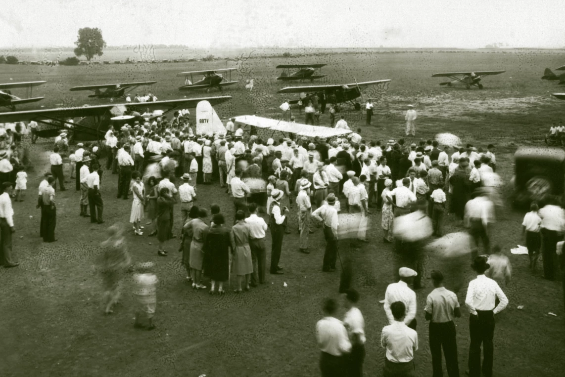 10 or more planes are lined up on the tarmac, and dozens of people mill about. It appears to be summer, no men are wearing jackets, some have their sleeves rolled up.