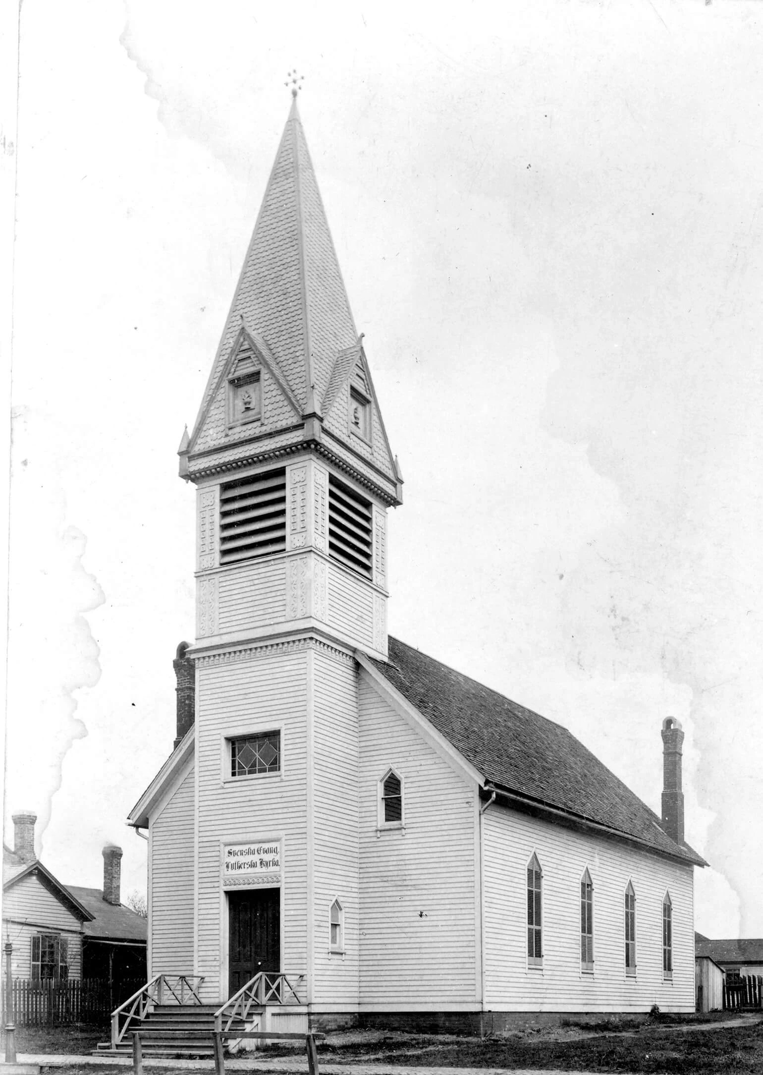 Black and white photo of a white church with wood siding and tall steeple in the front center.