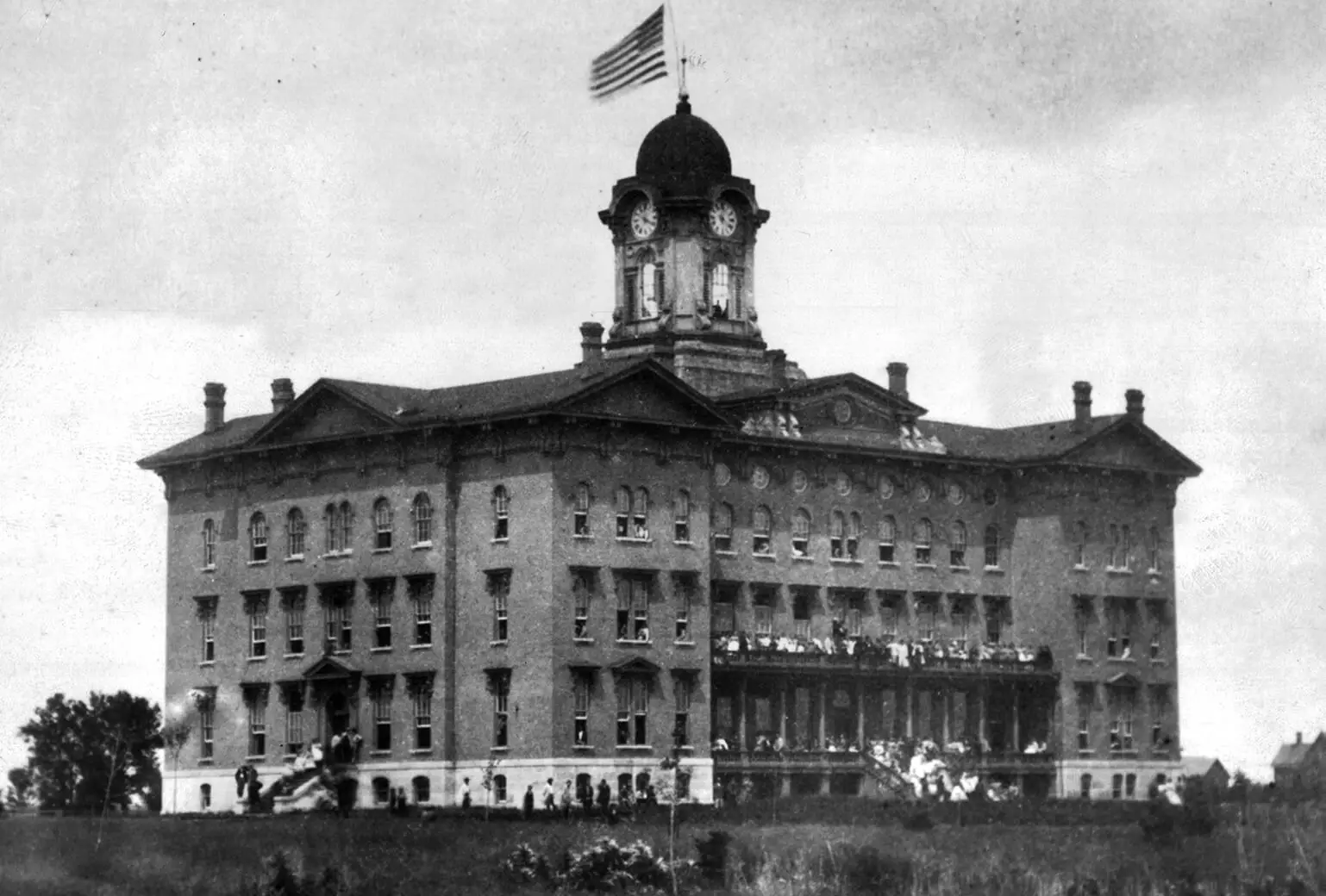 Black and white photo of a large four-story building with a central dome with clocks underneath it. The building design is square and very symmetrical, with a lot of windows.