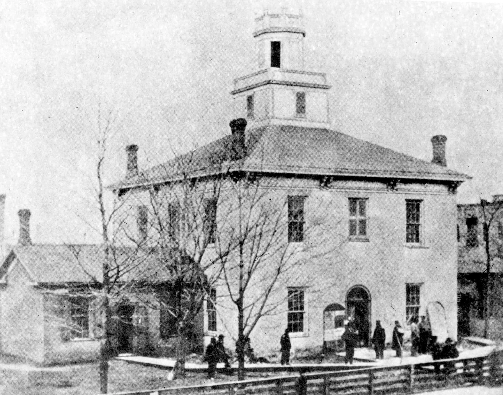 photo of a two-story building, the main part of the building is a square with a square cupula on top. on the first story a small addition sticks out from the side, where a sidewalk leads to the door.