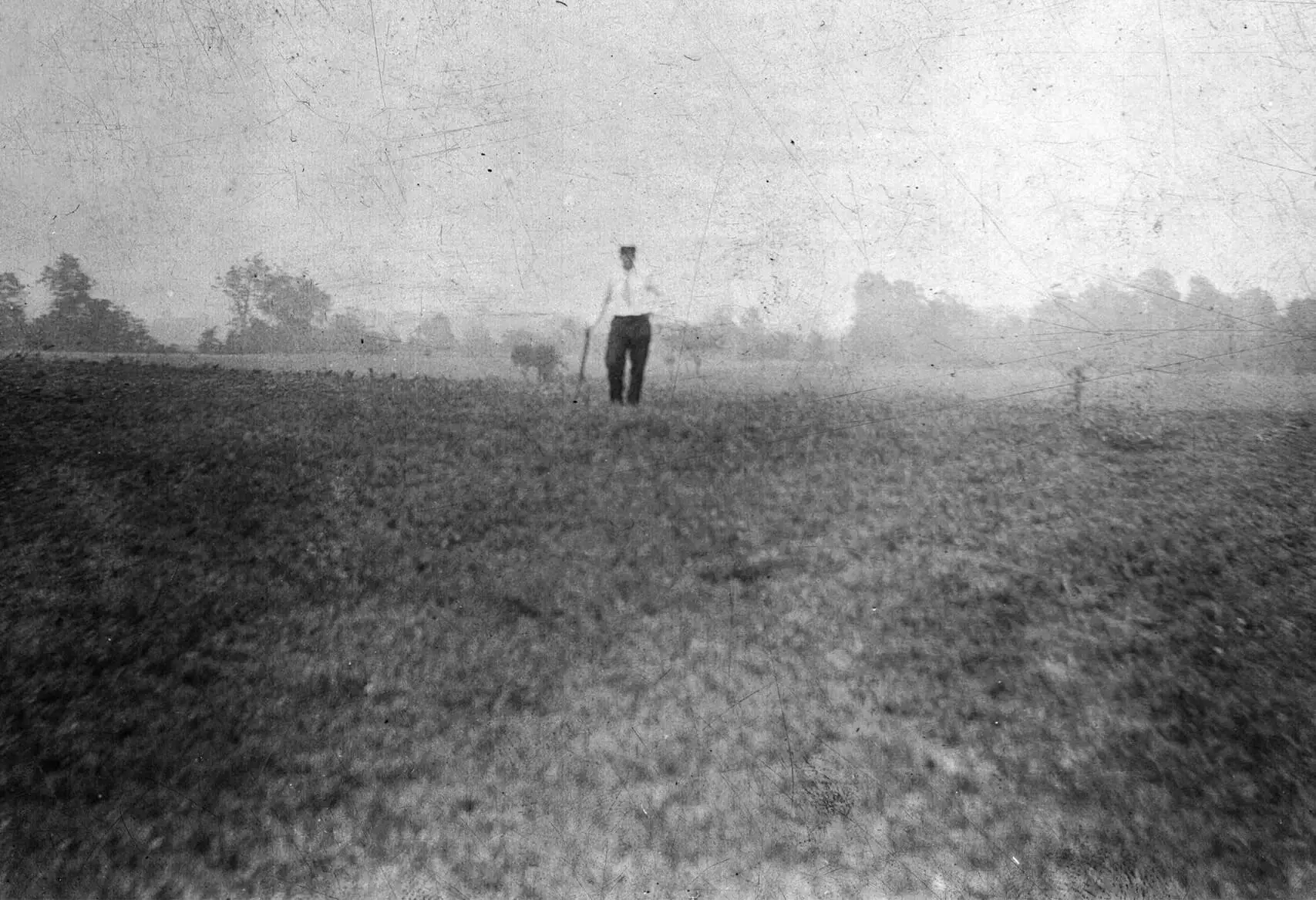 a man wearing a light shirt and dark pants stands in a field. he is leaning on a walking stick. there are trees in the distant horizon. The image is foggy, scratched, and not very focused.