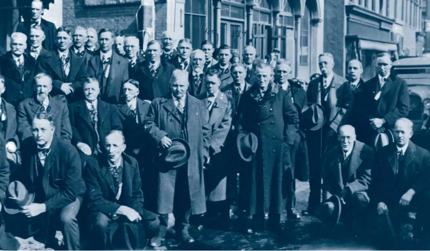 Group of 20-30 light-skinned men wearing suits, ties, and coats, holding their hats. None are smiling. They are standing outside on the sidewalk, overflowing into the street.