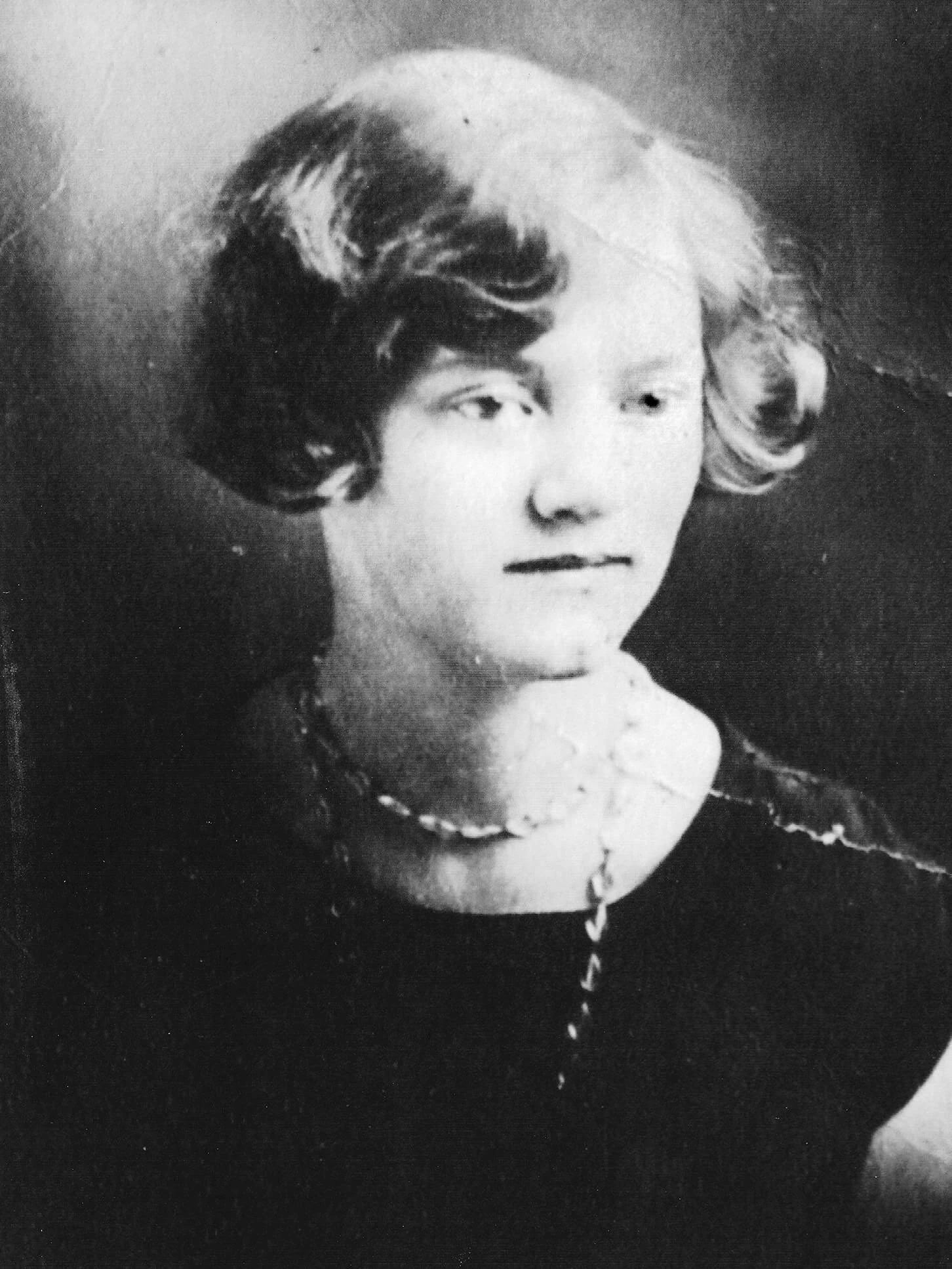 Black and white portrait of a young white woman with short dark hair. She is wearing a short sleeved dark shirt and has a long necklace wrapped around her neck