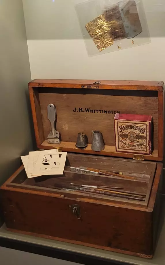 This wooden chest is open to reveal tools and items inside of it. There are brushes and scrapers and papers and a box of gold leaf. Above the box is a piece of gold leaf mounted with that brush that would be used to apply it. Gold leaf is an extremely thin layer of gold, so thin that a breath or breeze makes it flutter and break.