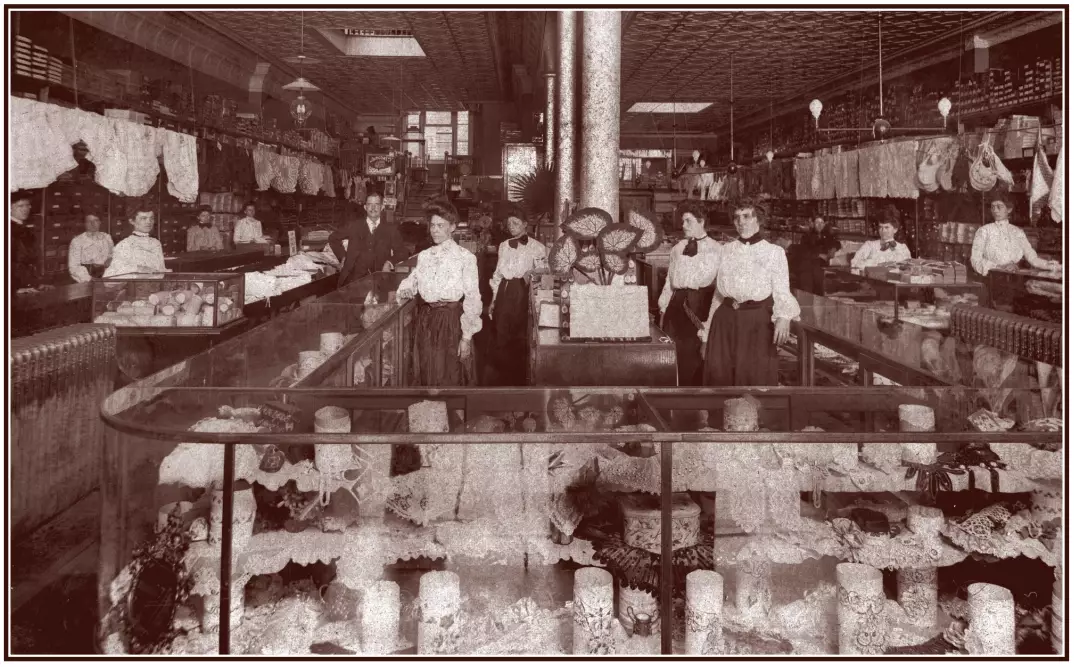 Portrait of women working at a clothing store. They are wearing white shirts and long dark dresses. They are surrounded by glass display cases and stacks of fabric on the back walls.