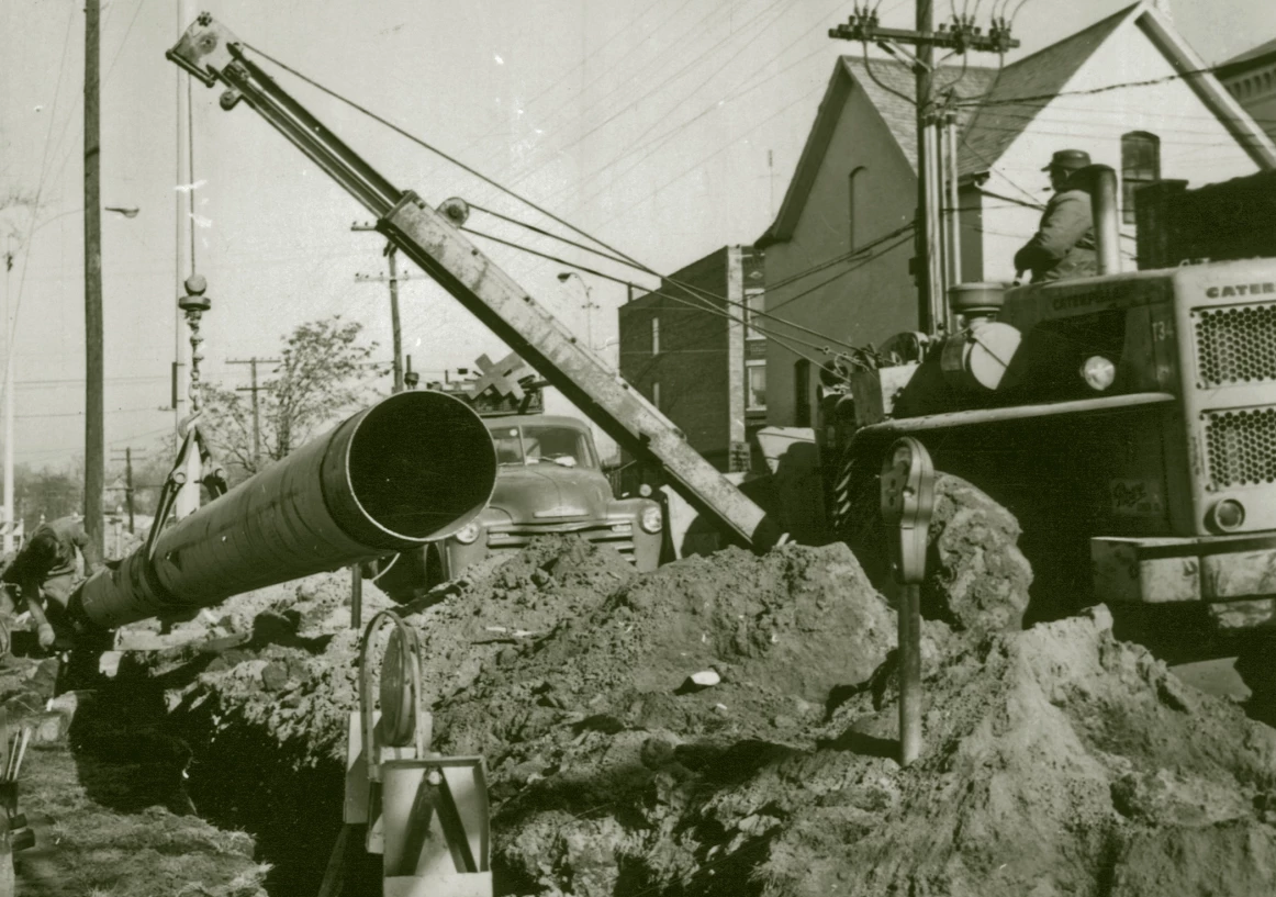 a small crane holds a large pipe, lowering into a ditch that was dug. They appear to be in town, there are buildings around, and a parking meter.