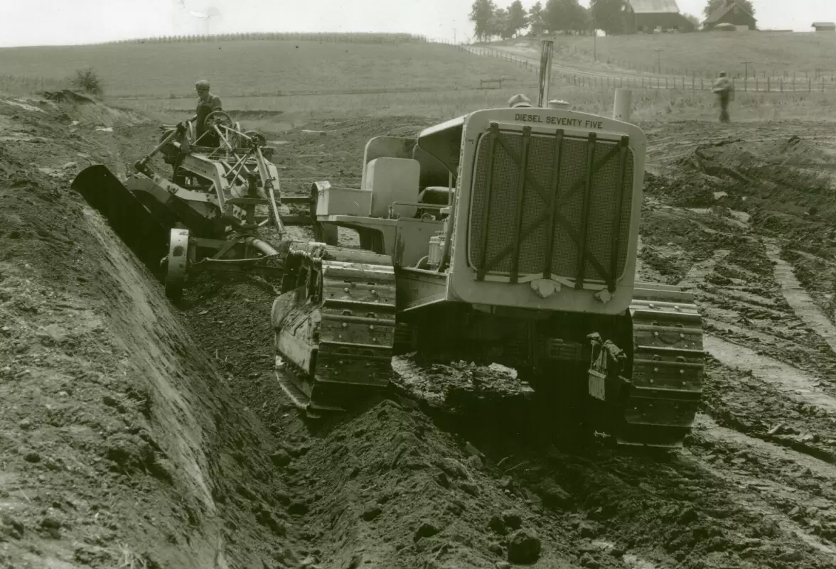 another image of the large tractor pulling a grader. This time they are surrounded by dirt and mud in the middle of a field.