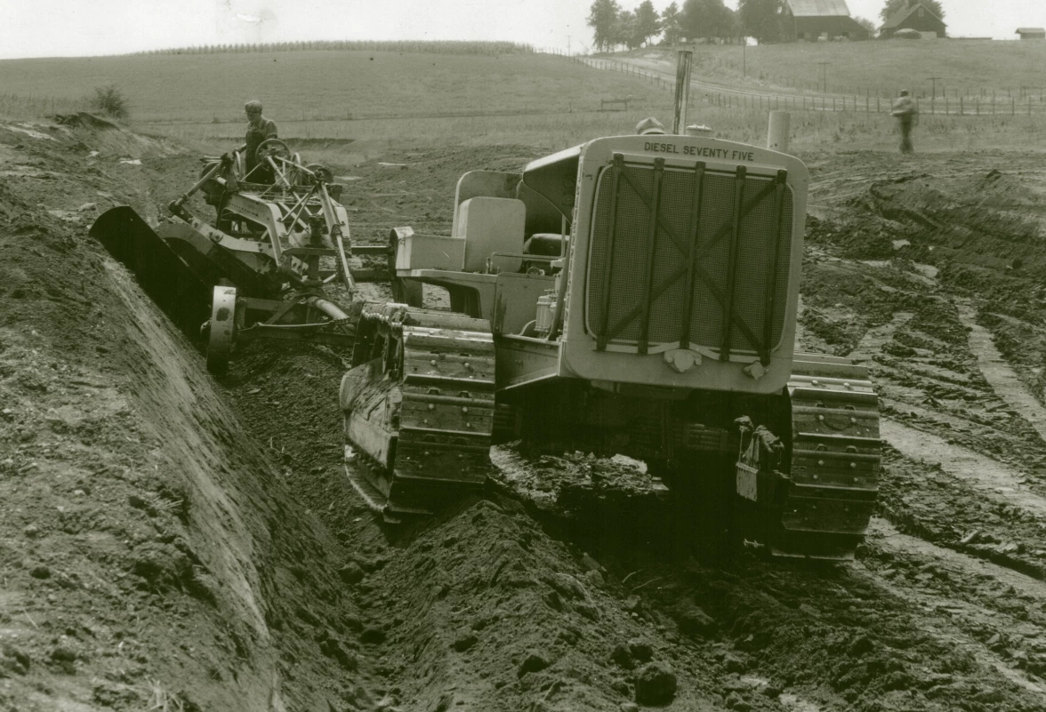 another image of the large tractor pulling a grader. This time they are surrounded by dirt and mud in the middle of a field.