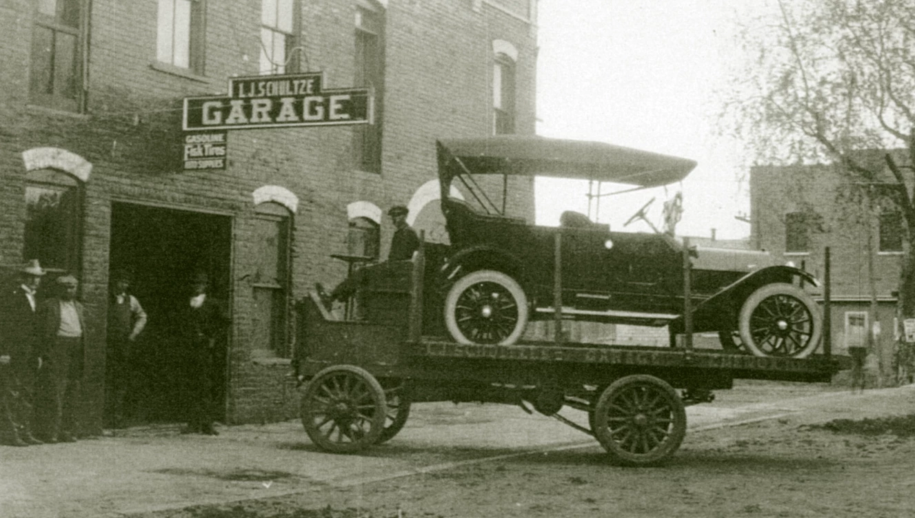 a flat bed truck with a brand new car on top drives towards a building.