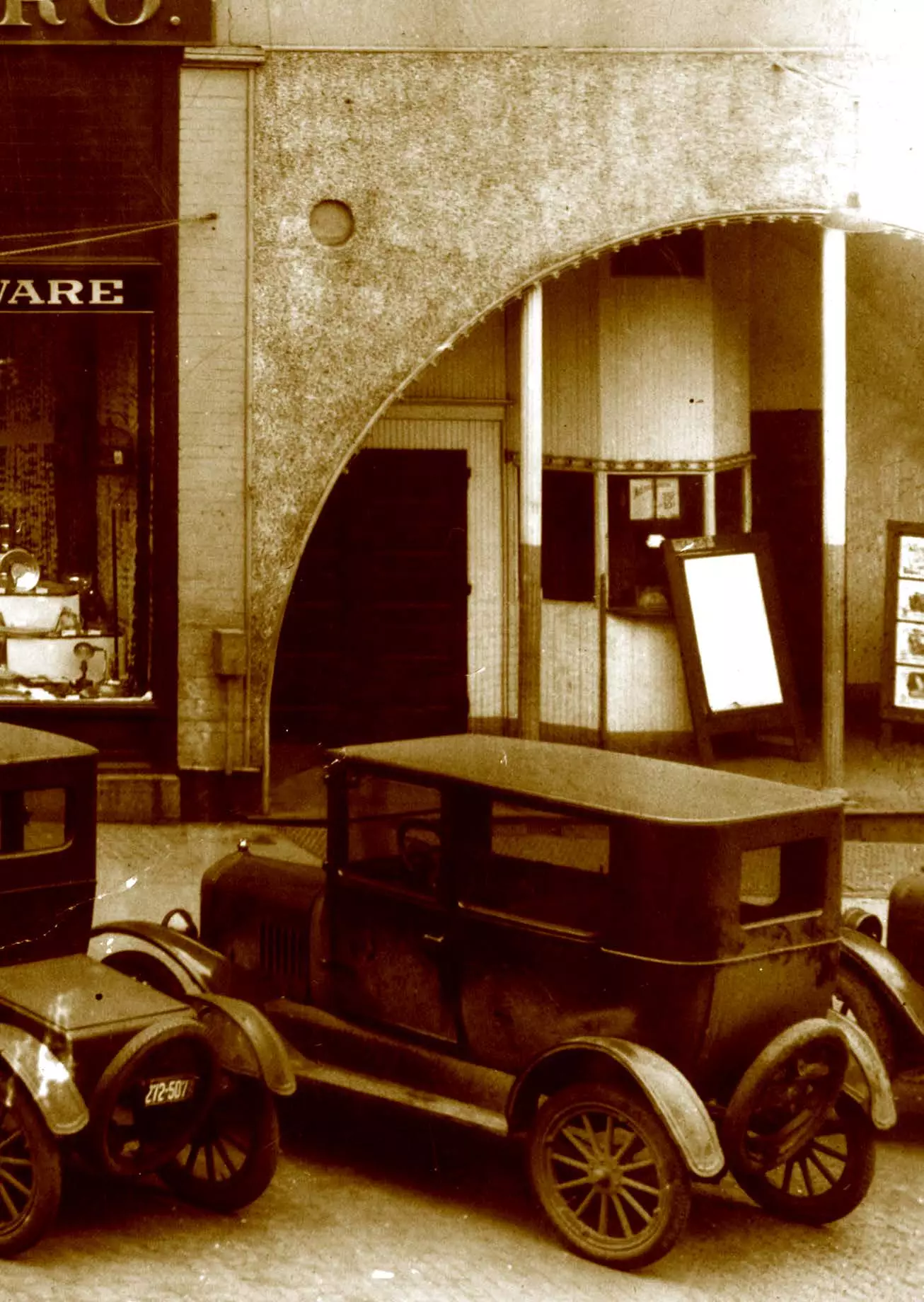 There are cars parked in the foreground. They are parked in front of a building that has an archway entrance. There are two supports holding up the archway. Is a walk-up window to purchase tickents. A sandwich board leans on that window, presumably that board would be set on the sidewalk when they are open.