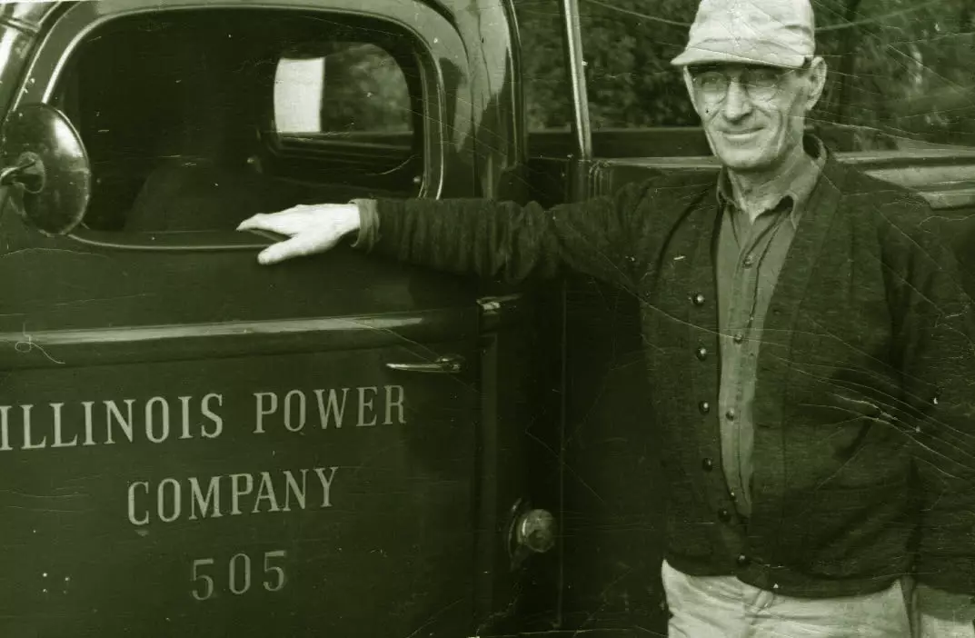 An older light-skinned man stands next to a truck. The window is down and he is resting his hand on the window opening.